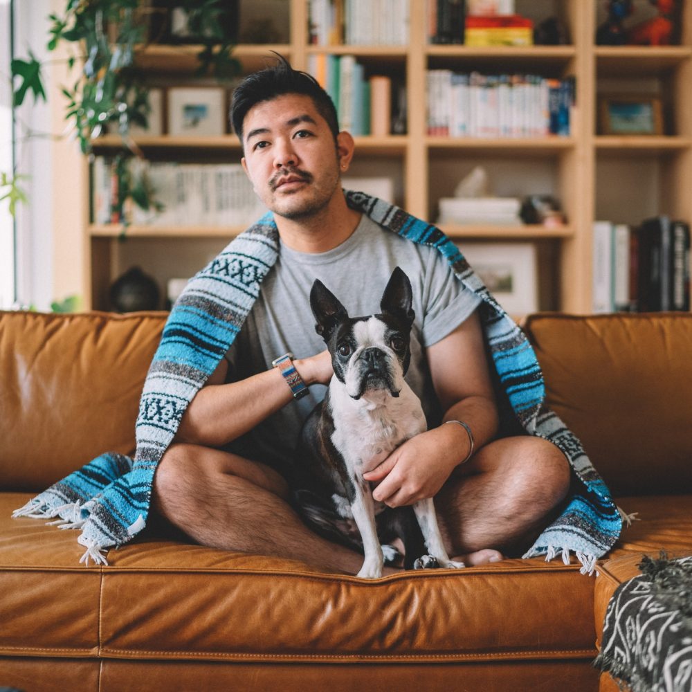 Guy sitting on couch, lounging with his dog watching TV with a blanket.